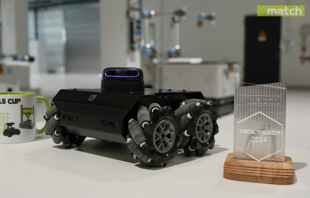 Ein robuster, sechsbeiniger Roboter mit einer Kamera auf der Oberseite steht auf einem Tisch neben einer Tasse mit der Aufschrift "DIGITAL TOOLS CUP" und einem klaren Acrylpokal, auf dem "INTEGRATION DIGITAL TOOLS CUP 2024" steht.